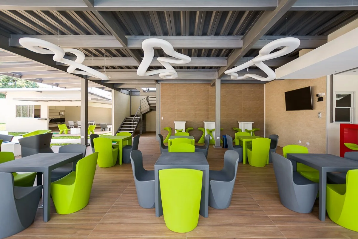 Modern outdoor lounge area with lime green and gray chairs and tables, a flat-screen TV on the wall, and abstract white ceiling lights, featuring a staircase leading to an upper level.