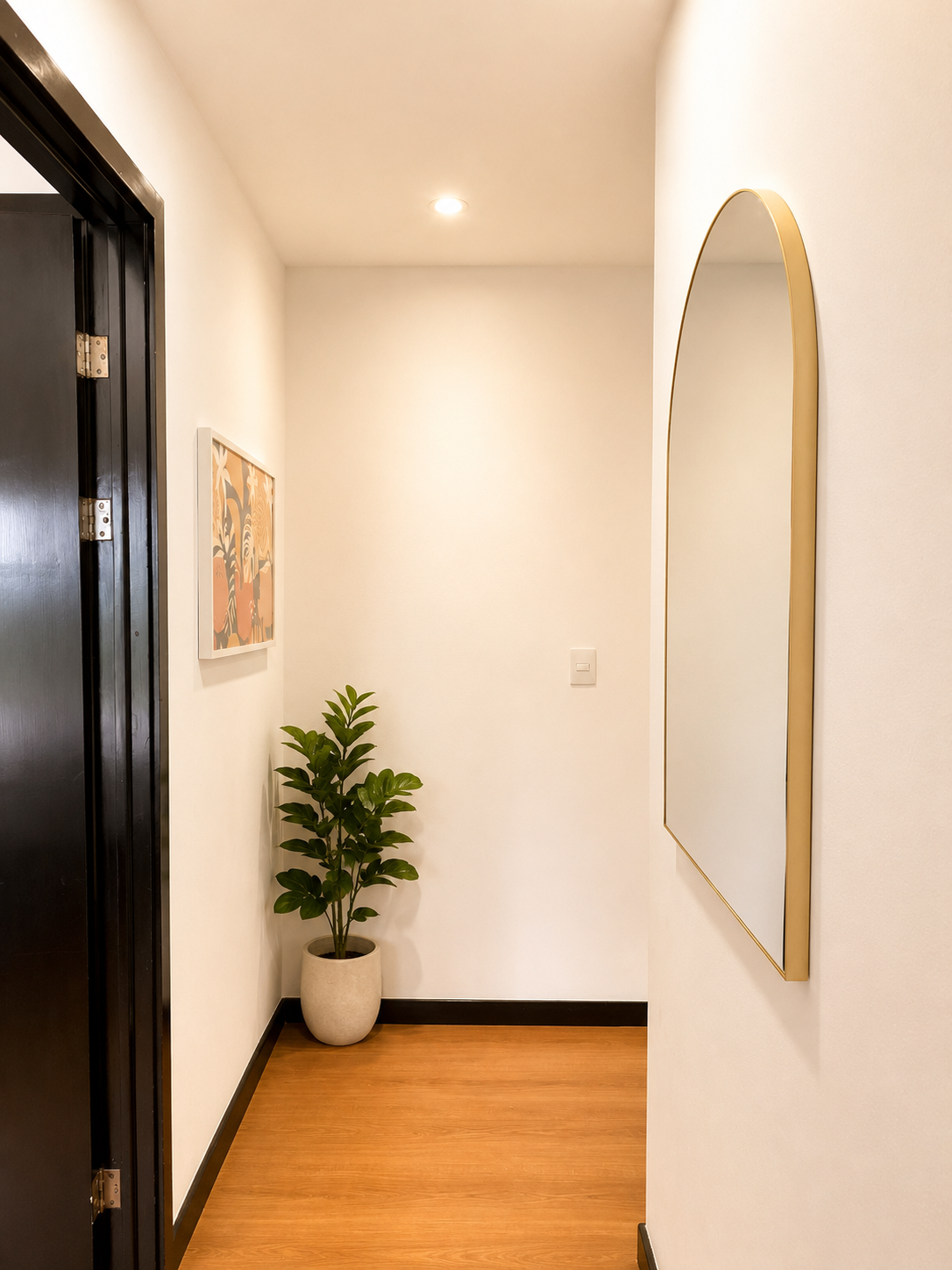 A narrow hallway with a wooden floor, a potted plant in a white planter, a framed artwork on the wall, and a tall oval mirror with a gold frame.