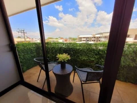Balcony with two chairs and a small round table with a yellow flower vase, overlooking a hedge and building under a partly cloudy sky.
