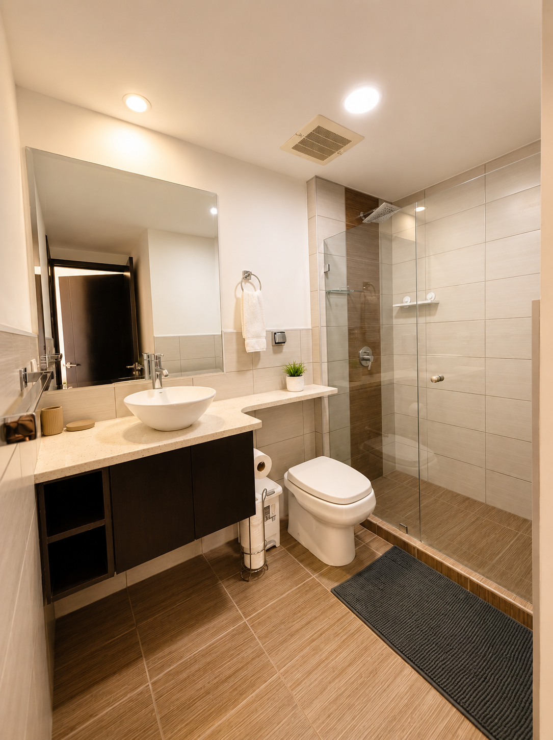 Modern bathroom with walk-in shower, white sink on beige countertop, large mirror, dark cabinet, toilet, towel, small plant, and black rug.