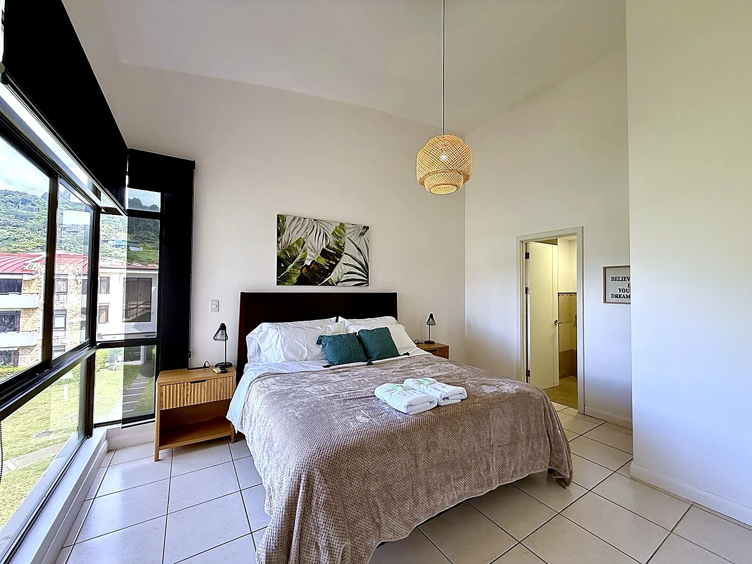 A neatly made bed with white and dark teal pillows in a bright hotel room with large windows, a wooden nightstand with lamps, and minimalist decor including a leaf print picture and a motivational quote on the wall.