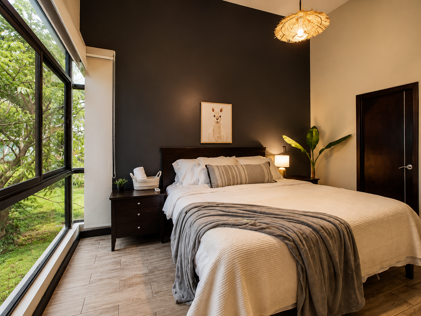 A bedroom with a large bed, dark wood headboard, white bedding, and a gray throw blanket. A striped gray pillow rests on the bed. There is a nightstand with a small plant, basket, and lamp. A framed llama picture hangs above the bed. A large plant is