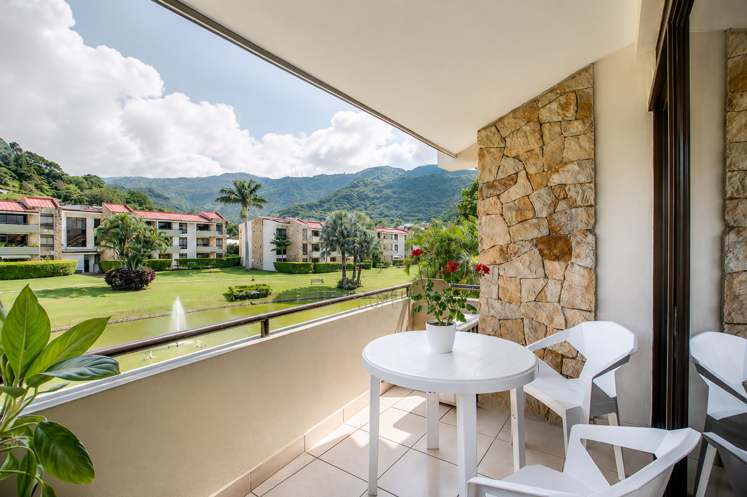 Balcony view of a lush green landscape with apartment buildings, palm trees, and mountains in the background, featuring a white table with potted red flowers and white chairs.