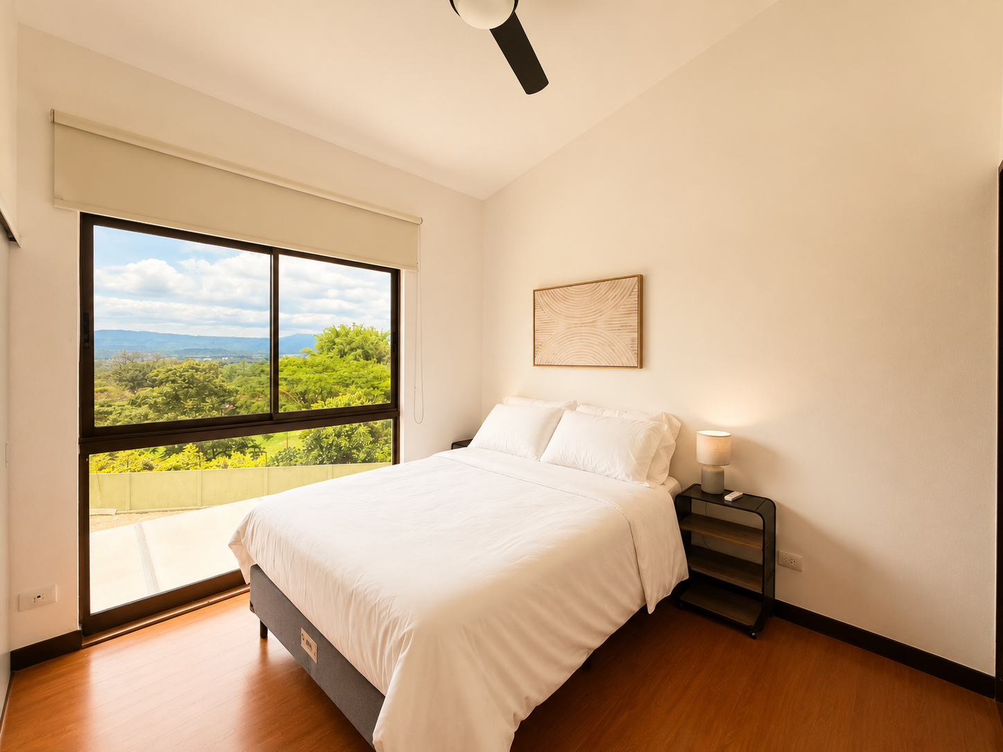 A bedroom with a large window showing a scenic view of mountains and trees, a white bed with pillows, a small nightstand with a lamp, and a wall art piece above the bed.