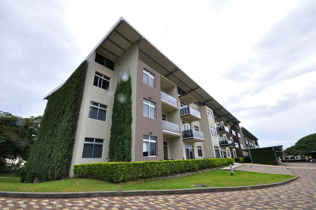 A modern multi-story residential apartment building with balconies, partially covered in green ivy, and surrounded by a paved driveway and well-maintained lawn.