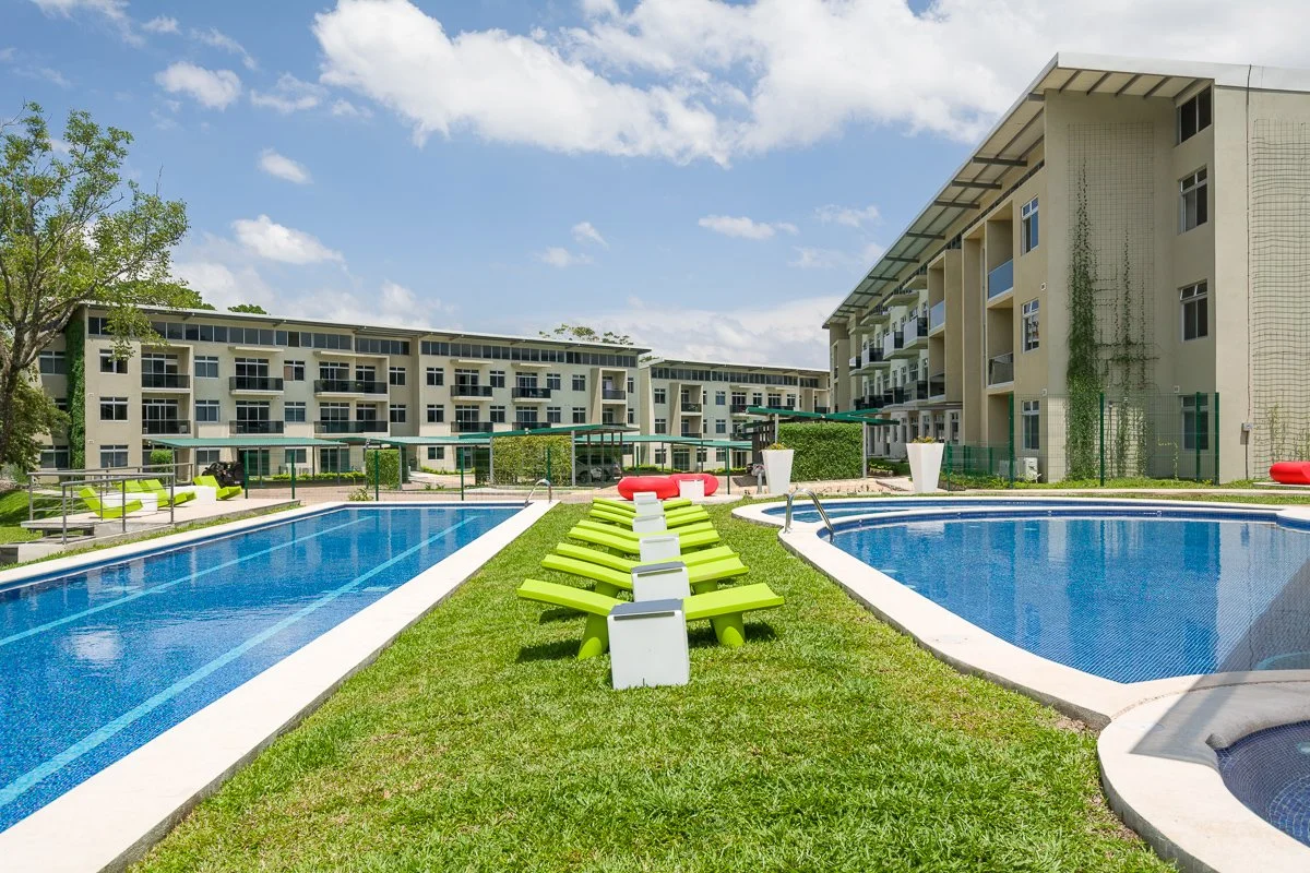 Apartment complex with two swimming pools, green lounge chairs, and modern buildings under a partly cloudy sky.