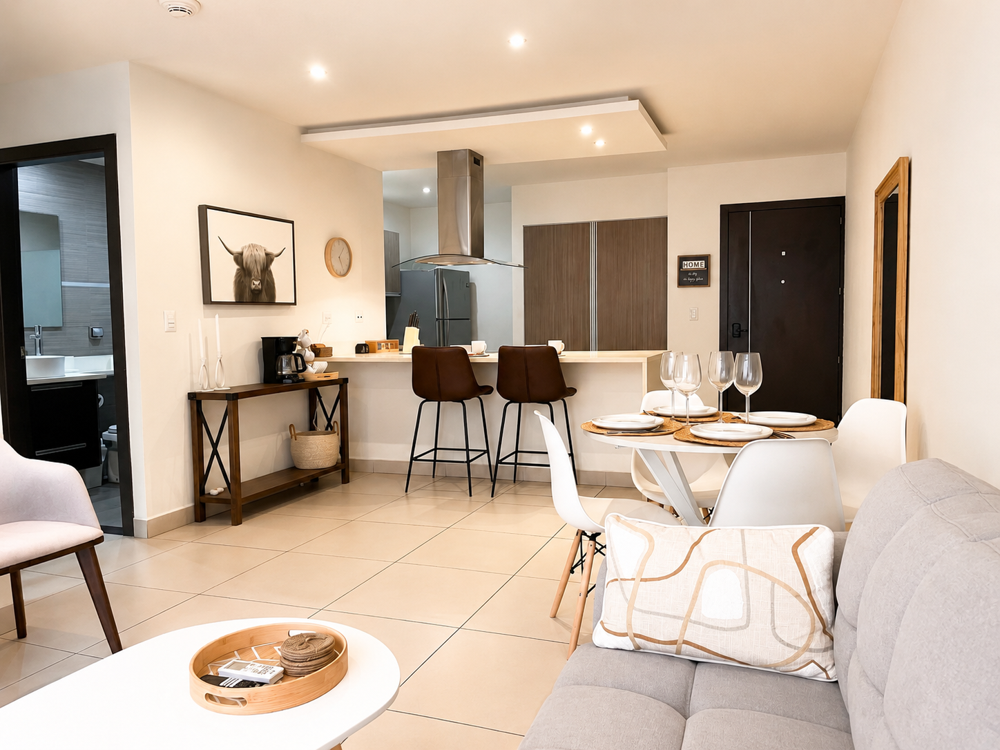 Open-concept living and dining area with a kitchen in the background. The living space has a light gray sofa with a decorative pillow, a white coffee table with a wooden tray, and beige tiled floors. The dining area features a round white table set w