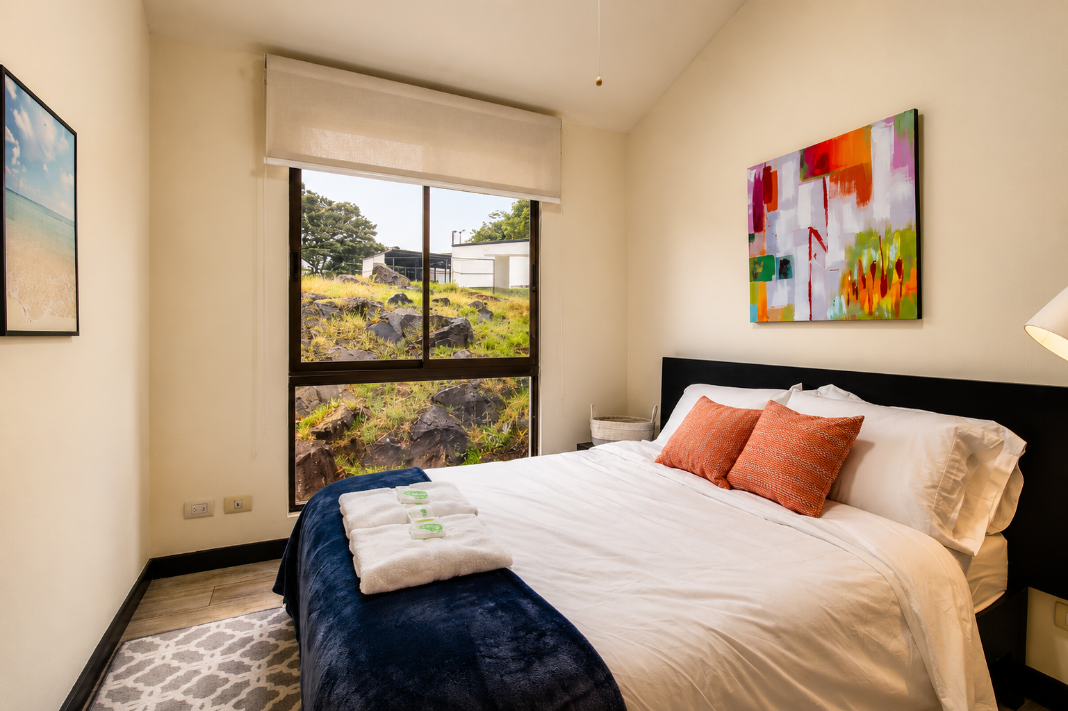 A tidy bedroom with a made bed featuring white and orange pillows, a dark headboard, and a navy blue throw. There is a window showing a rocky and grassy outdoor scene, with colorful abstract artwork on the wall. Towels and soap are on the bed, and a 
