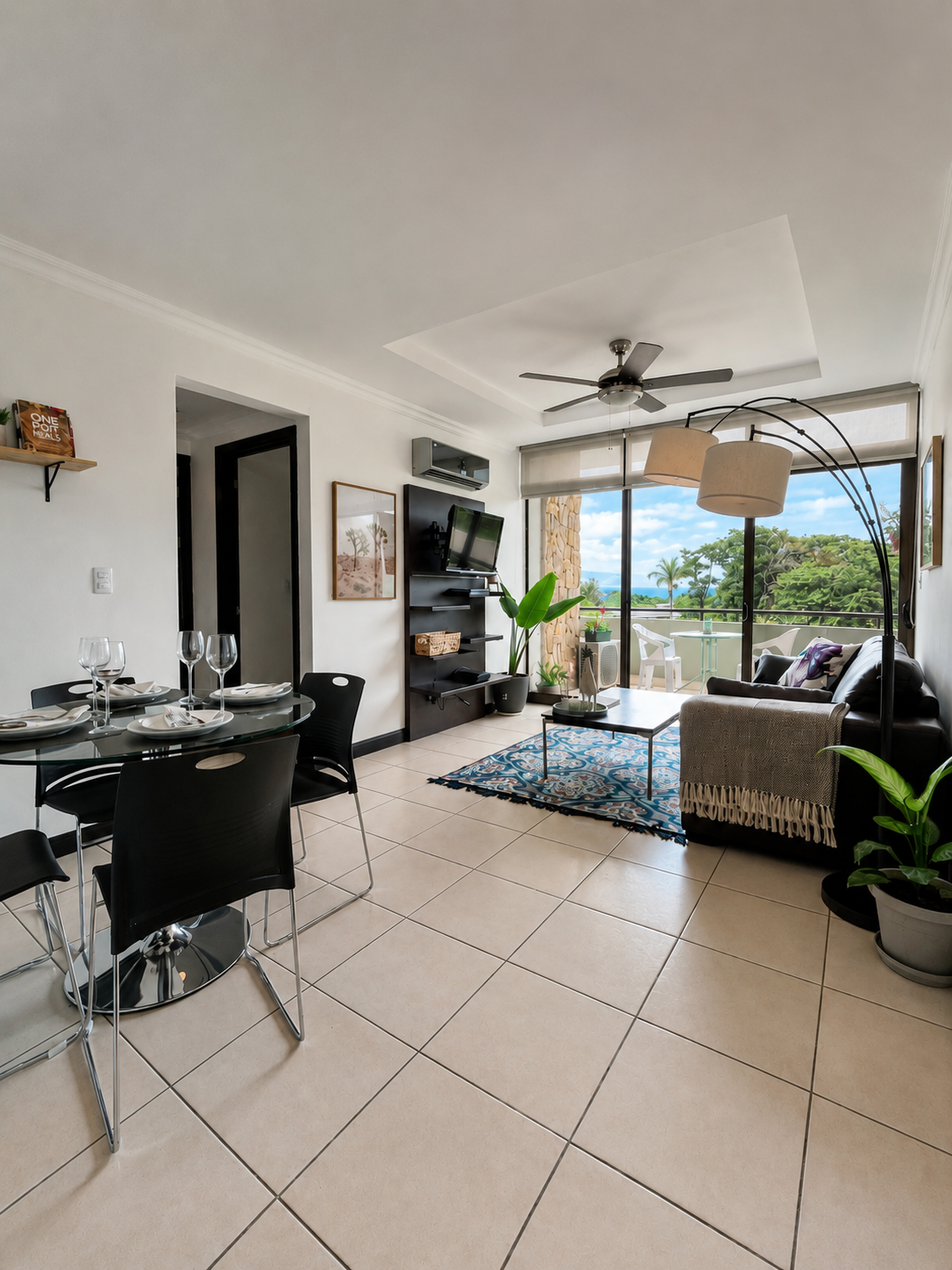 Living room and dining area in an apartment with open sliding glass door leading to balcony, featuring a black couch, potted plants, a glass-top dining table with black chairs, a TV mounted on a black shelf, and a ceiling fan.