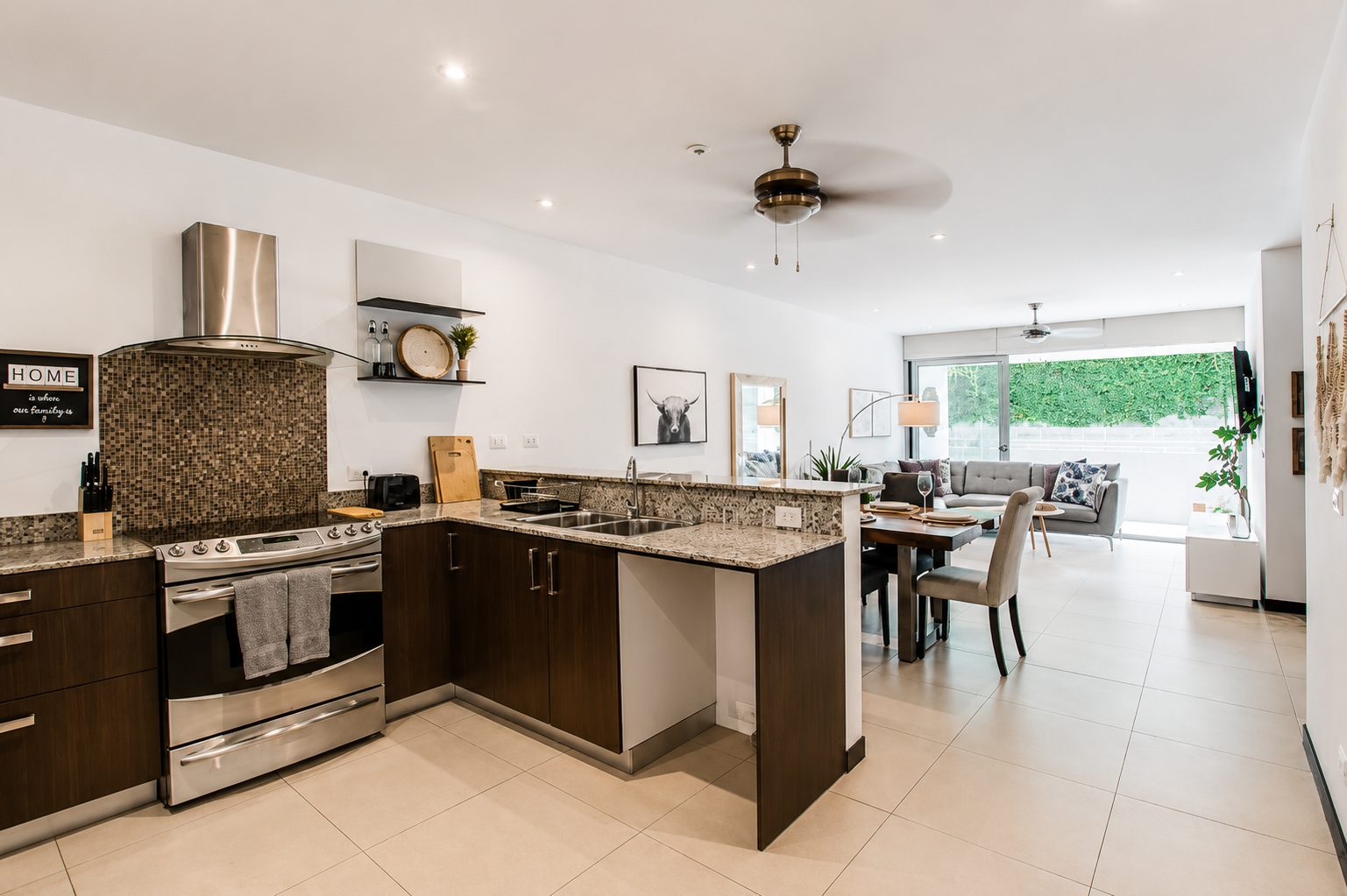 Open-concept kitchen and living room with modern decor, granite countertops, stainless steel appliances, dining table with chairs, and a large window with greenery outside.