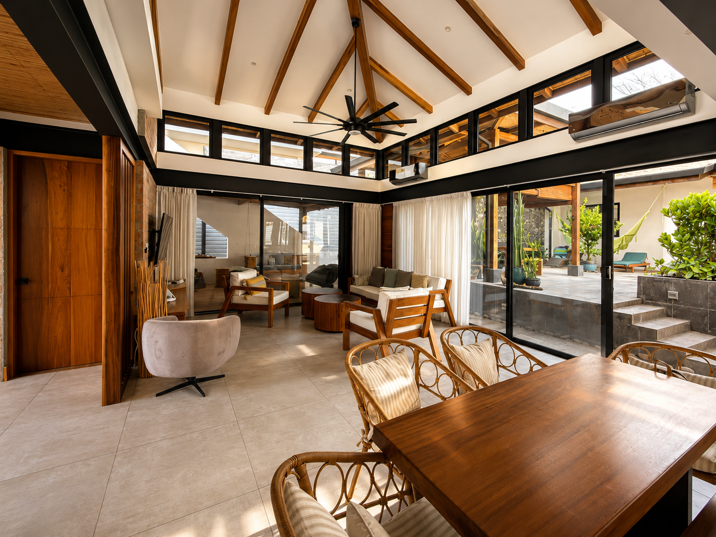 Living room with high ceiling, wooden beams, ceiling fan, glass sliding doors, indoor plants, and modern furniture including sofas, chairs, and a dining table.