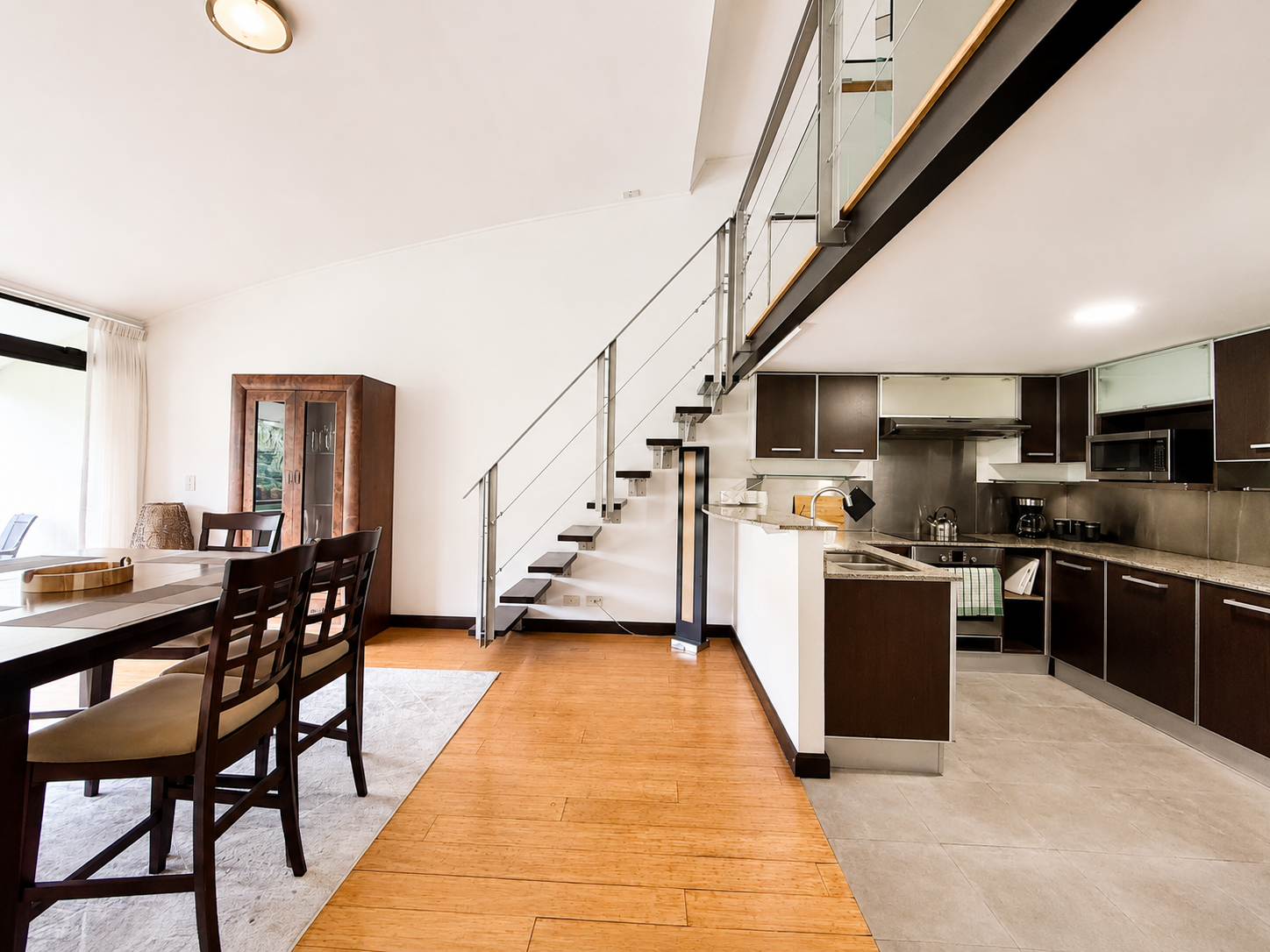 Open-plan living area with a dining table, chairs, a wooden cabinet, a kitchen with dark cabinets, stainless steel appliances, and a staircase with metal railing.