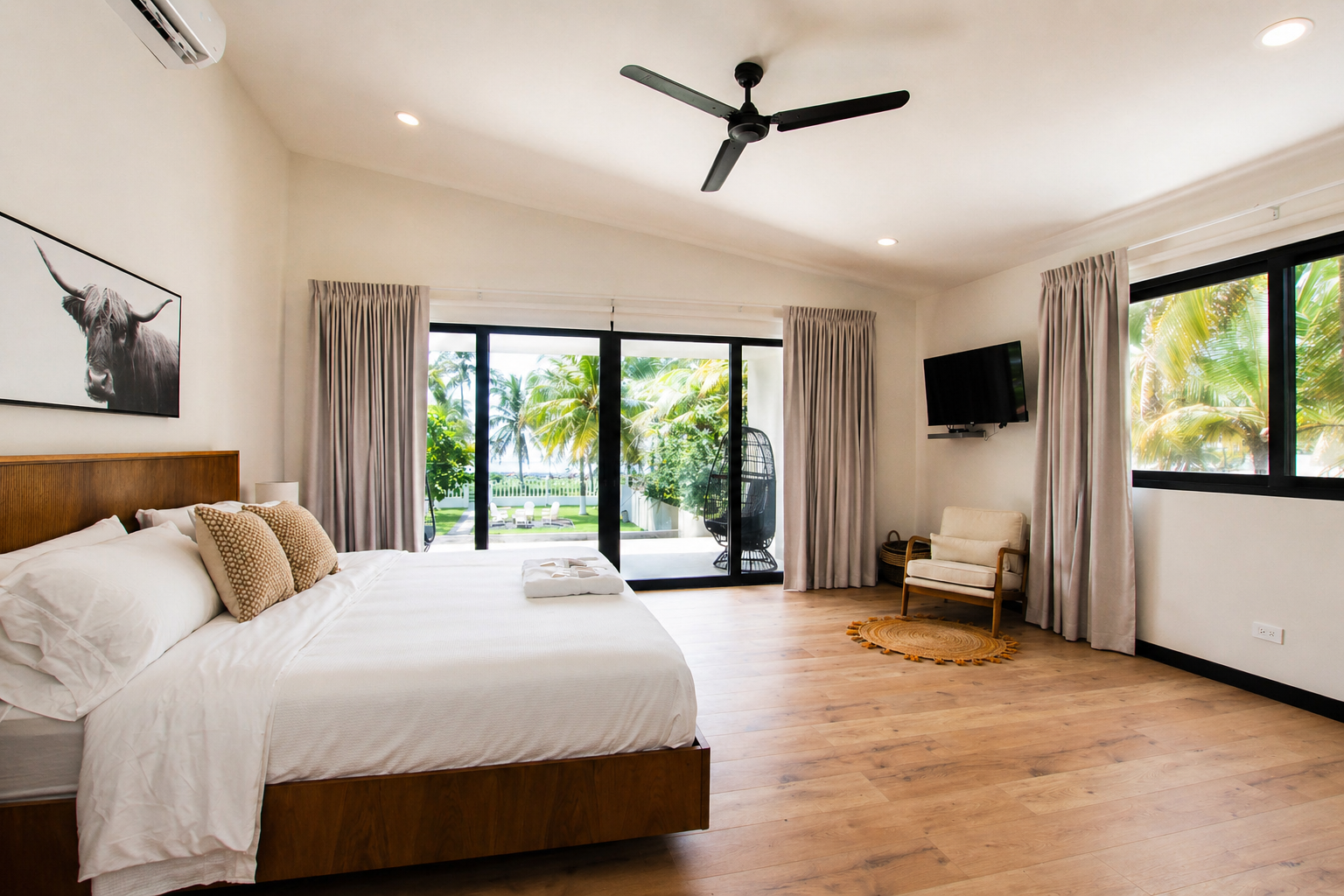 Spacious bedroom with a large bed, wooden headboard, and bedside lamps. There's a balcony with glass doors and tropical greenery outside. The room has a ceiling fan, a wall-mounted TV, and a sitting area with a chair and small rug. Beige curtains fra