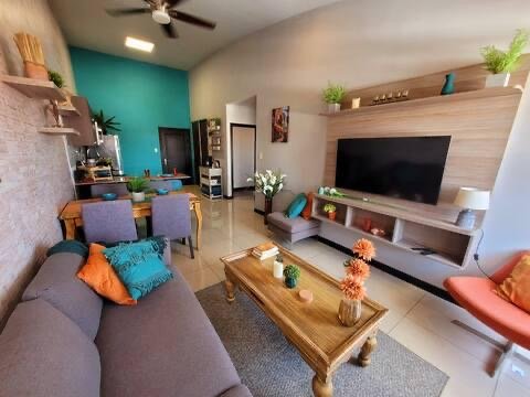 Living room with a gray sofa, orange and blue pillows, a wooden coffee table, a TV mounted on a wall unit with shelves, potted plants, and a dining area with six chairs in the background.
