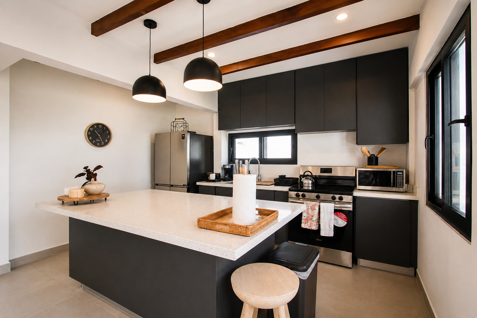 Modern kitchen with black cabinets and white quartz countertops, a central island with a paper towel roll, black pendant lights, a stainless steel refrigerator, oven, microwave, and various kitchen utensils, with a window providing natural light.