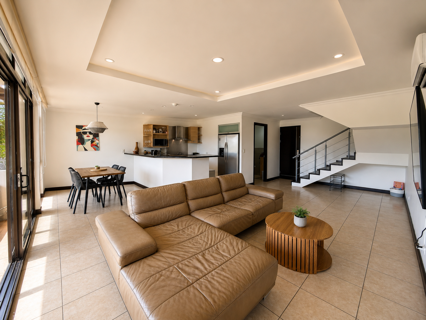 Living room with brown leather sectional sofa, wooden coffee table with a potted plant, open kitchen with black counter, dining table with black chairs, staircase with metal railing, sliding glass doors to the patio, and art on the wall.