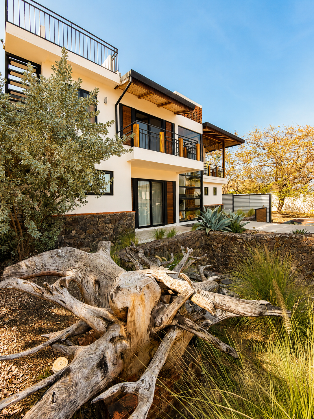 Modern multi-story house with white exterior walls, large windows, and multiple balconies. The house features a stone foundation and is surrounded by desert landscaping with succulents, agave, and driftwood, under a clear blue sky.
