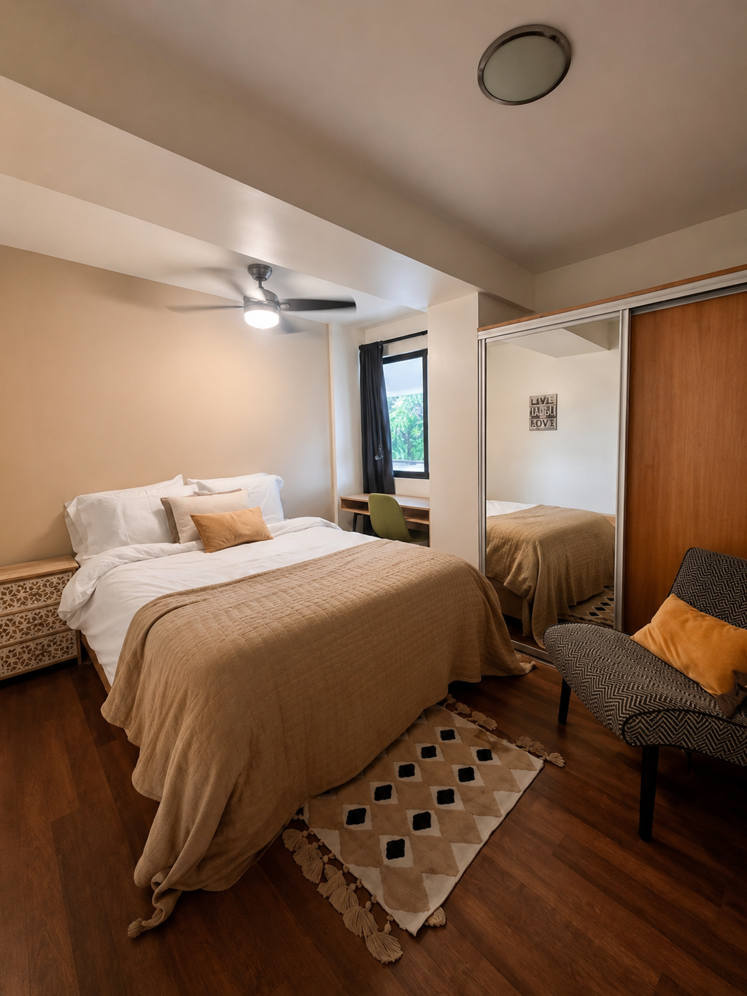 A cozy bedroom with a bed covered in a beige blanket, white pillows, and a small tan pillow. There is a patterned rug at the foot of the bed. A black ceiling fan with a light is above, and a window with black curtains is on the side. There is a small