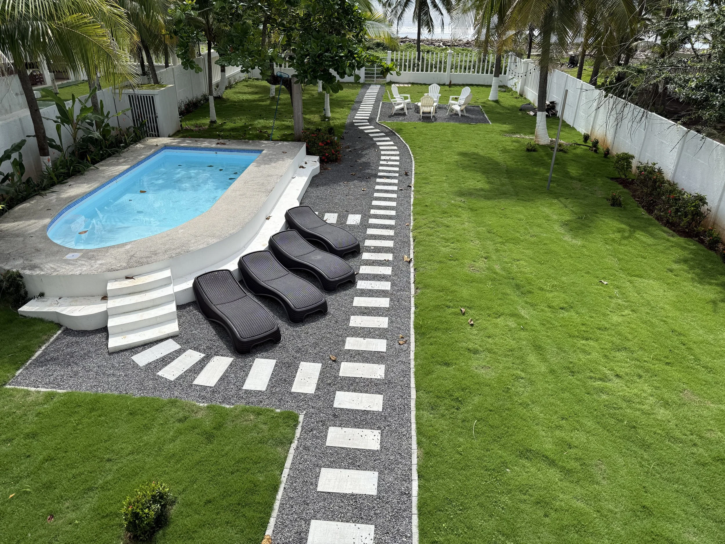 A backyard with a small swimming pool, a paved walkway with black and white tiles, and a sitting area with five white chairs and a table. The yard is enclosed with a white fence and features green grass, trees, and bushes.