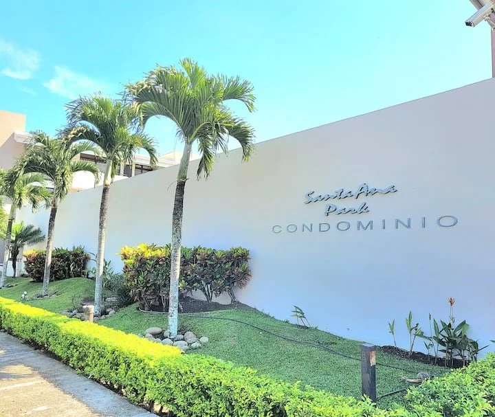 Exterior view of Sultana Park Condominio with palm trees, green hedges, and a white wall with the condominium's name.