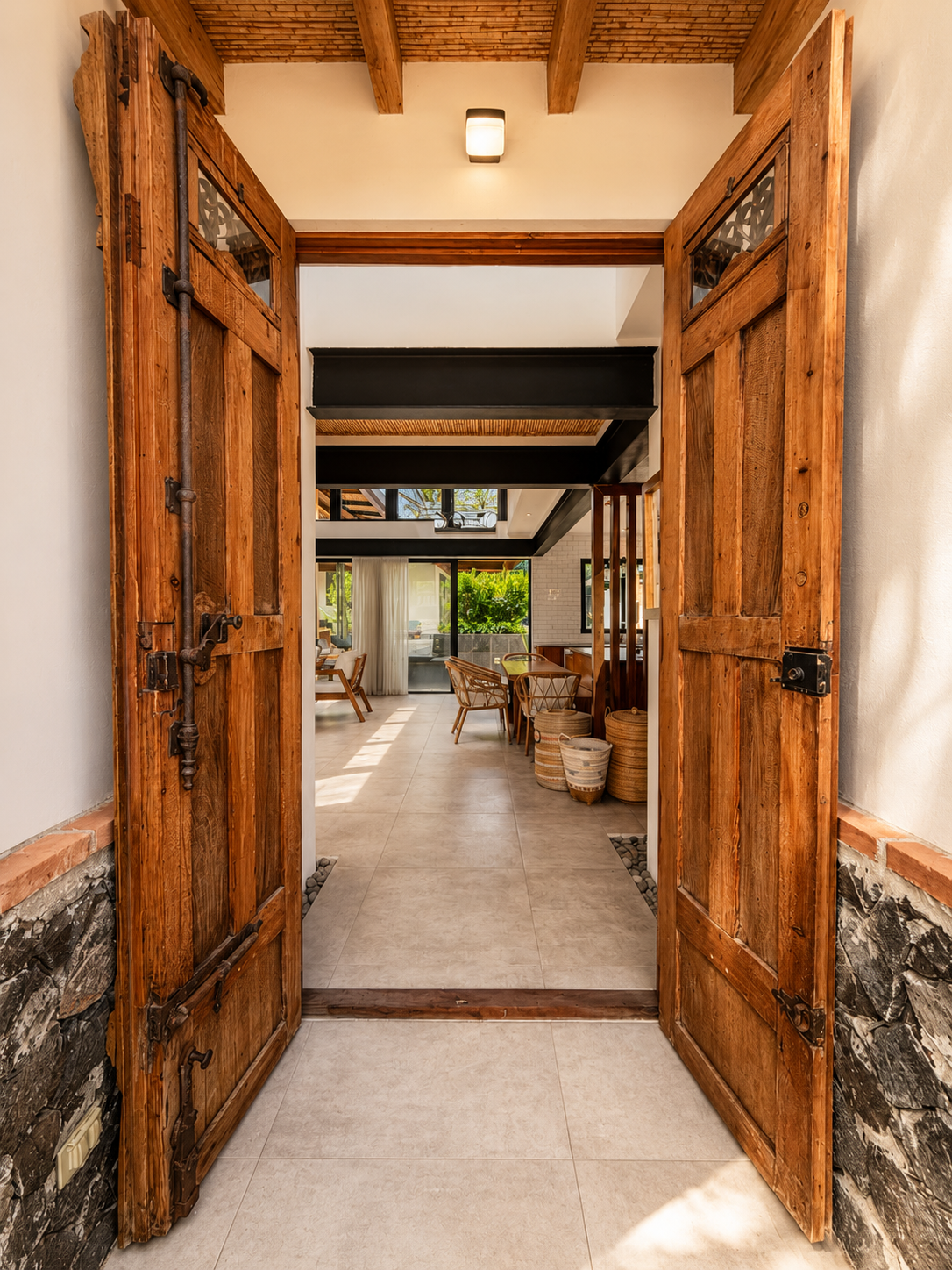 Wooden double doors open to a modern interior with a dining area and large windows showing outdoor greenery.