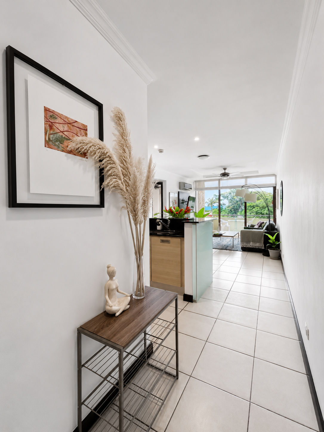 Interior view of a modern apartment with white walls, tile flooring, and minimalist decor. A console table with a decorative sculpture and tall dried pampas grass is placed along the wall. In the background, there's a kitchen with a black countertop,
