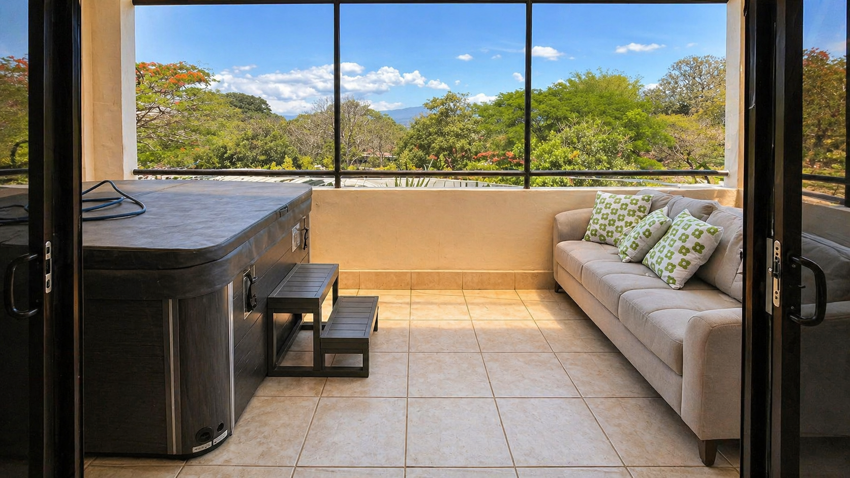 Balcony with a hot tub, a beige sofa with green and white patterned pillows, and large windows showing a view of trees and a blue sky.