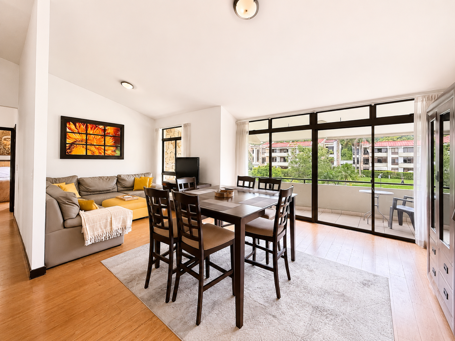 Bright living room with a dining table, chairs, a sofa with yellow cushions, wall art, and large windows with a balcony view of greenery and buildings outside.