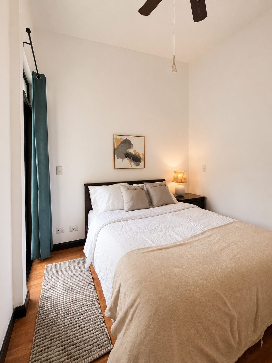 Clean, minimal bedroom with a bed, nightstand, lamp, artwork, and a ceiling fan.