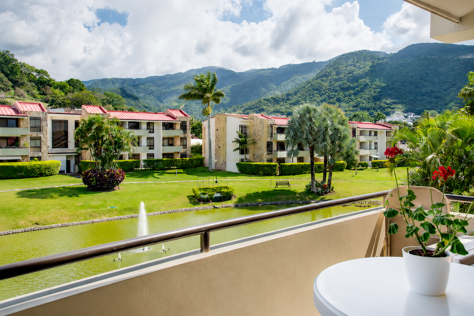 View from a balcony overlooking a landscaped garden with a water fountain, palm trees, bushes, and multi-story apartment buildings with red roofs and stone accents, with mountains in the background and partly cloudy sky.