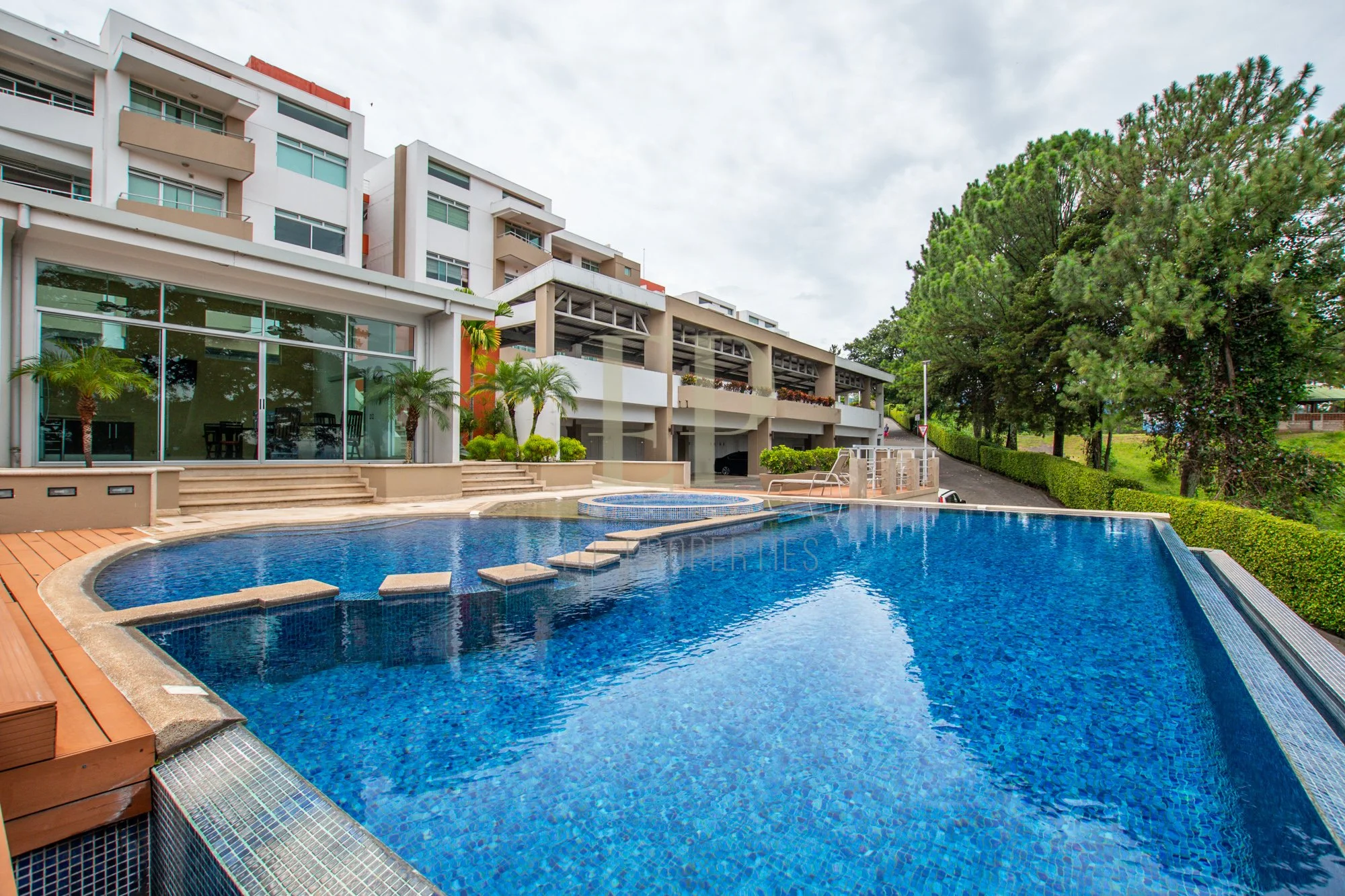 A modern residential building with balconies and large glass windows, situated next to a swimming pool with stepping stones and a surrounding deck with outdoor furniture, greenery, and trees under a cloudy sky.