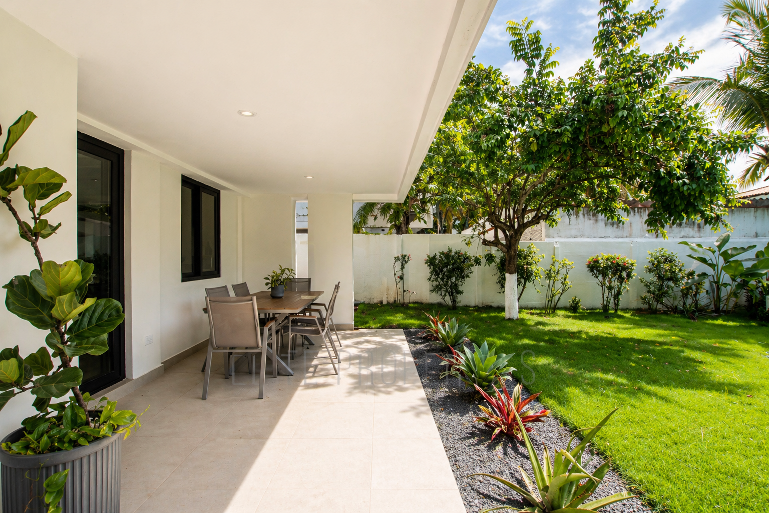A covered patio area with outdoor dining table and chairs next to a garden with green grass, plants, and trees, in a backyard setting.