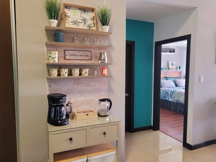 Kitchen nook with coffee station, including a coffee maker, an electric kettle, and cups, with shelves above holding plants, glasses, and decorative items, adjacent to a bedroom with a bed and tropical-themed bedding.