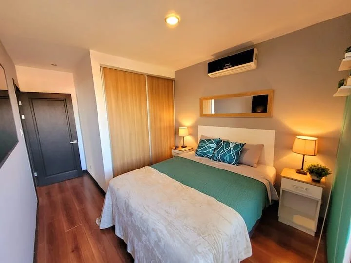 A cozy bedroom with a bed, blue pillows, and a green blanket. There are two nightstands with lamps and a small plant. A mirror is above the bed, and a split air conditioner is mounted on the wall. The room has a wood floor and a closet with sliding d