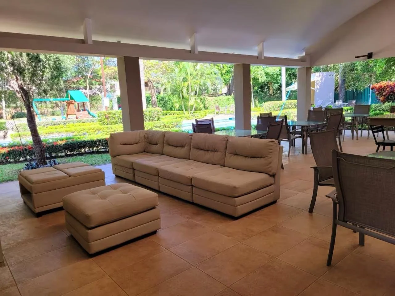 Indoor seating area with a beige sofa, matching ottomans, and multiple tables and chairs. Large glass windows reveal a backyard with green trees, a lawn, and a playground with a slide.