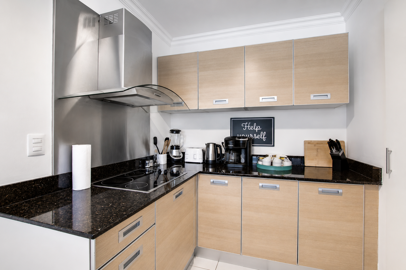 Modern kitchen with beige cabinets, black granite countertops, and stainless steel appliances, including a range hood and coffee maker, with a small 'Help yourself' sign on the wall.
