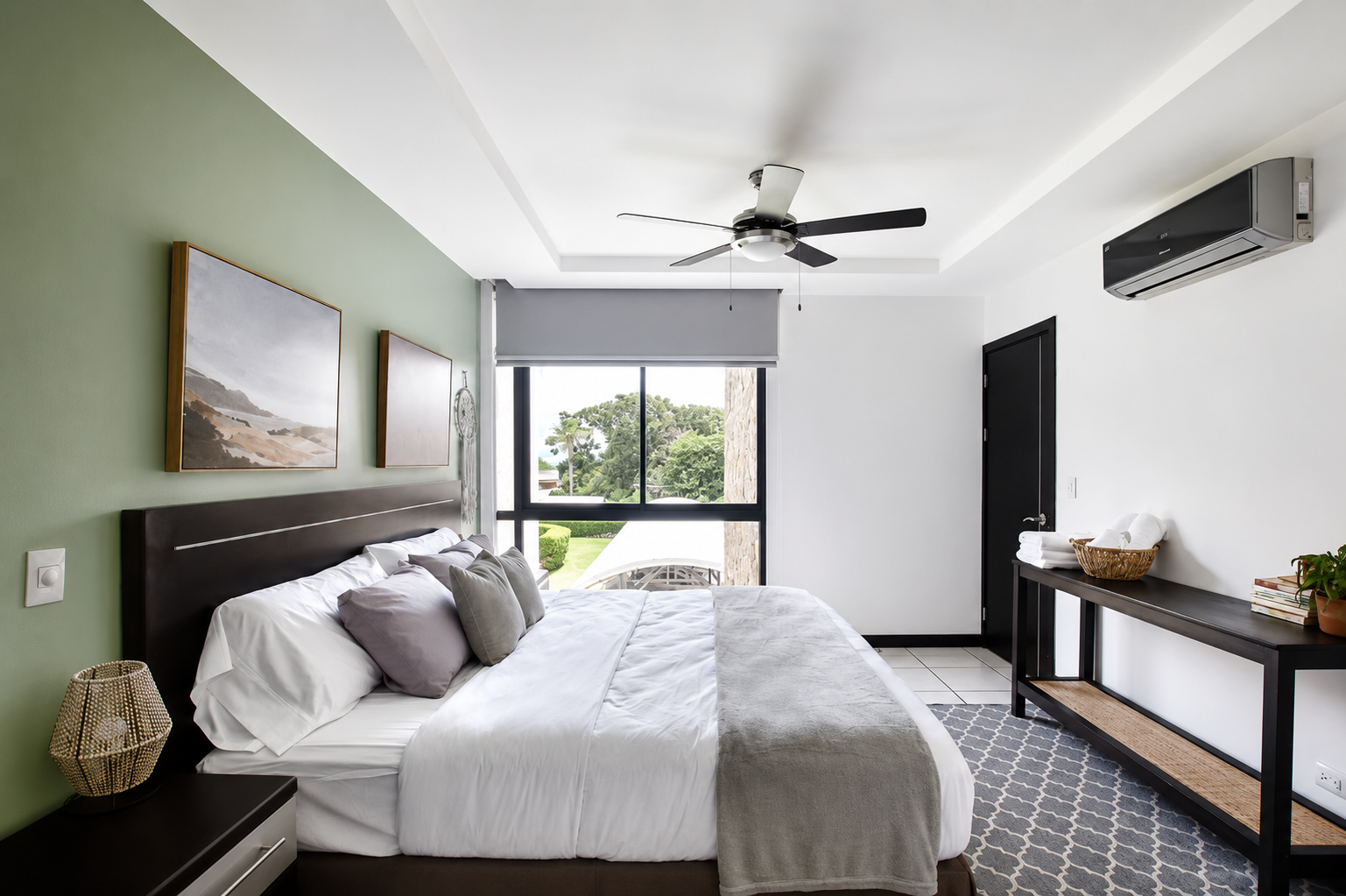 Modern bedroom with a large bed, decorative pillows, artwork on the green wall, a bedside lamp, black door, window with view of greenery, ceiling fan, air conditioning unit, and a black console table with towels, books, and a potted plant.