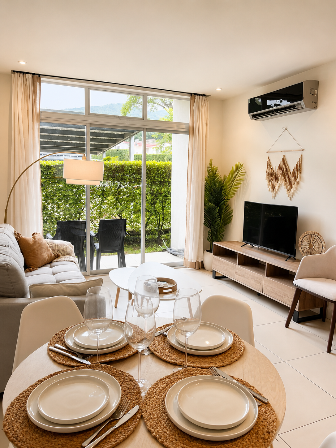 Living room with patio view, dining table set with plates and glasses, TV and decorative plants.