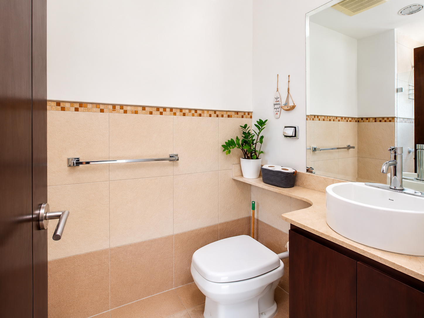 A clean bathroom with beige tiled walls, a white toilet, a wooden cabinet, a beige countertop with a round white vessel sink, a large mirror, a potted plant, and decorative wall hangings.