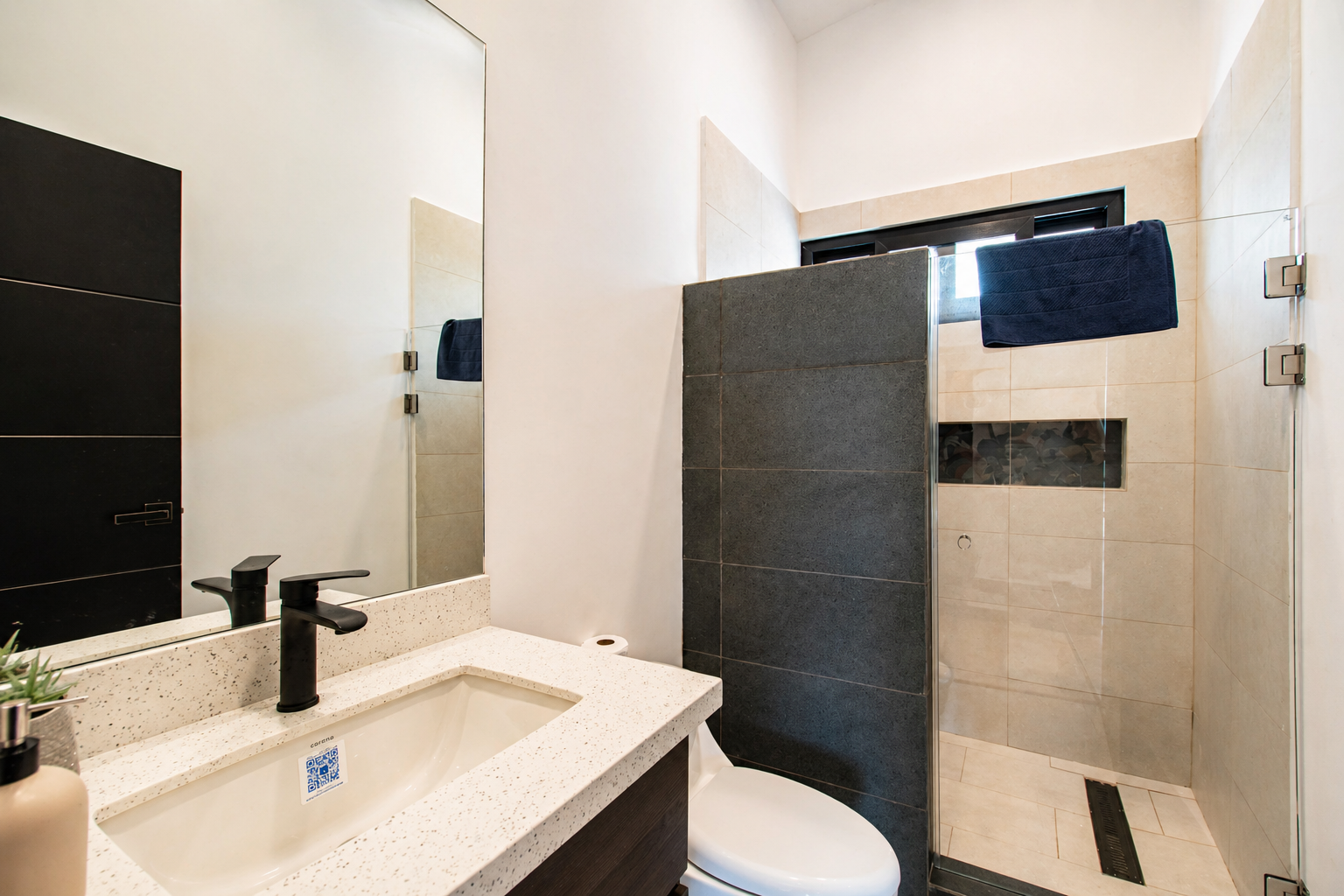 Small modern bathroom with white walls, black faucet, beige speckled countertop, mirror, and a toilet. There is a glass shower area with dark tiles and a small window with a towel hanging on a rack.