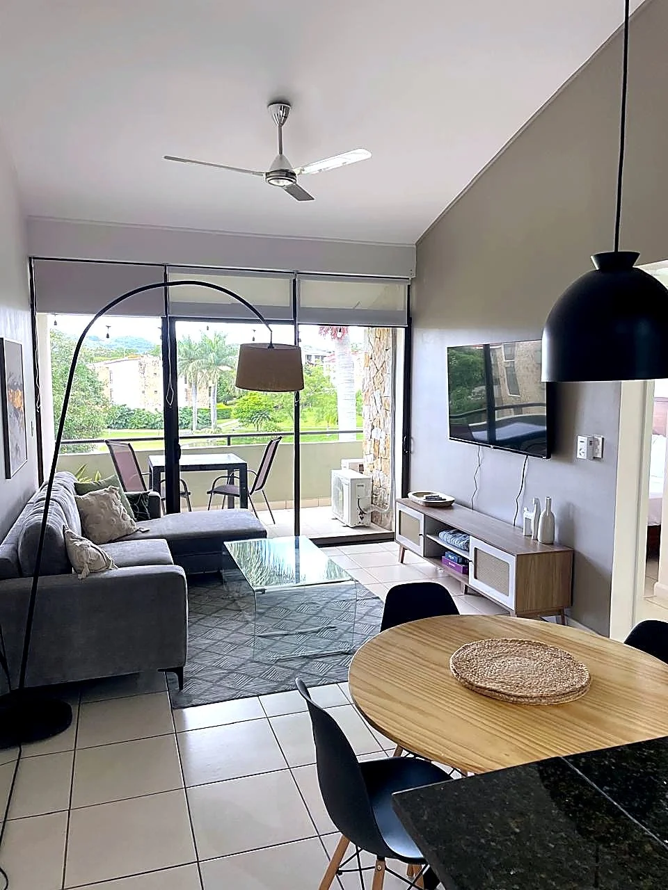 Living room with a gray sofa, glass coffee table, TV on the wall, dining table with black chairs, floor lamp, and balcony with outdoor seating view.