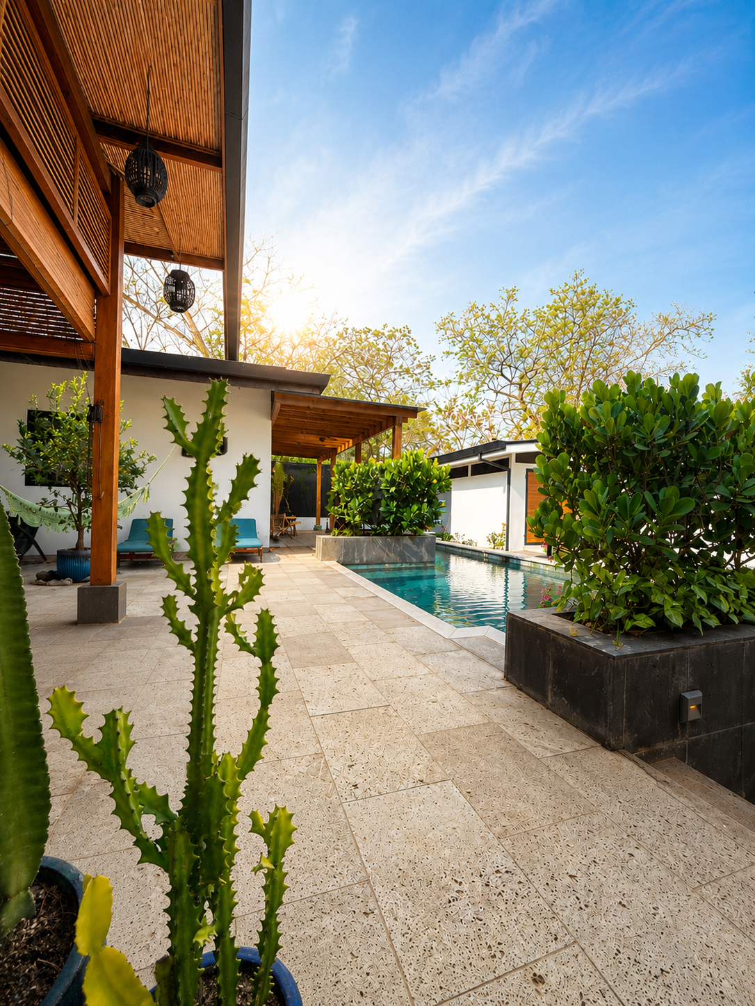 A modern backyard with a swimming pool, potted cacti, green bushes, and a wooden covered patio area under a blue sky with some clouds.