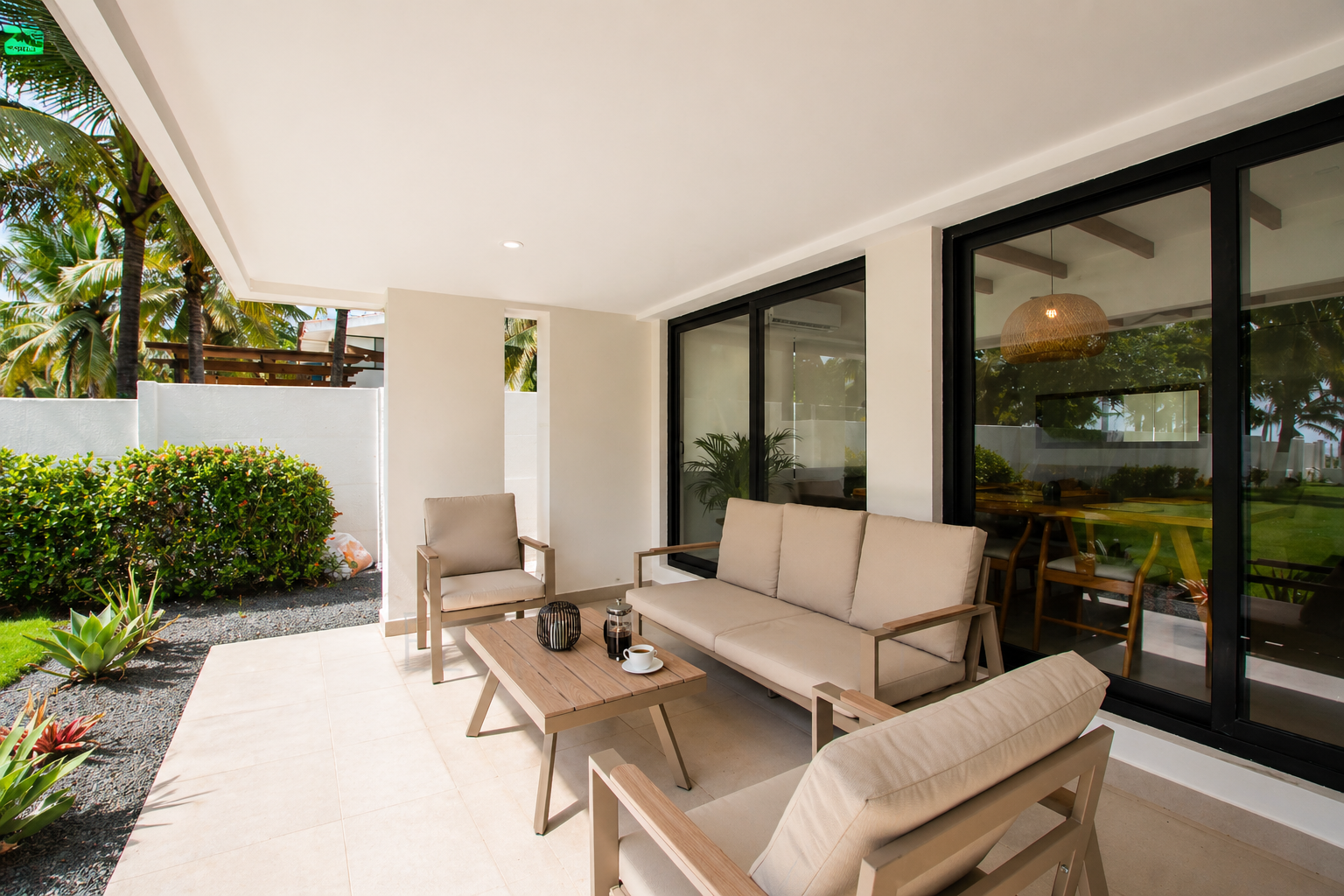 Outdoor patio with beige cushioned seating, wooden coffee table, potted plants, and glass sliding doors leading to an indoor dining area, surrounded by tropical greenery.