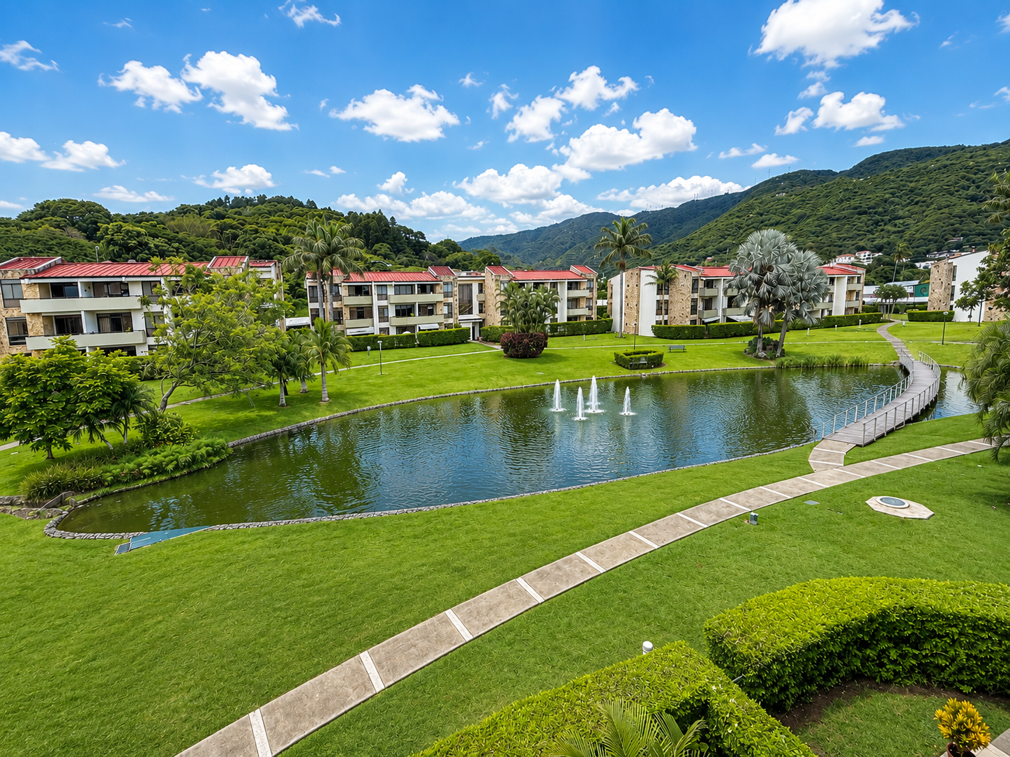 A landscaped residential complex with a pond, fountain, walking paths, green lawns, trees, and hillside in the background under a partly cloudy sky.