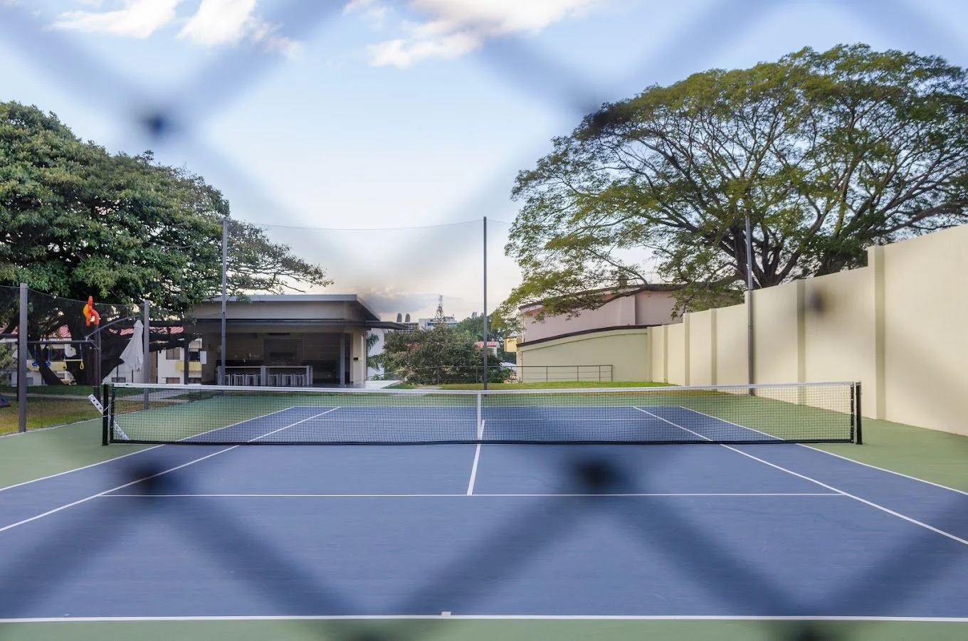 An outdoor tennis court viewed through a chain-link fence with a net, surrounded by trees and a building in the background.