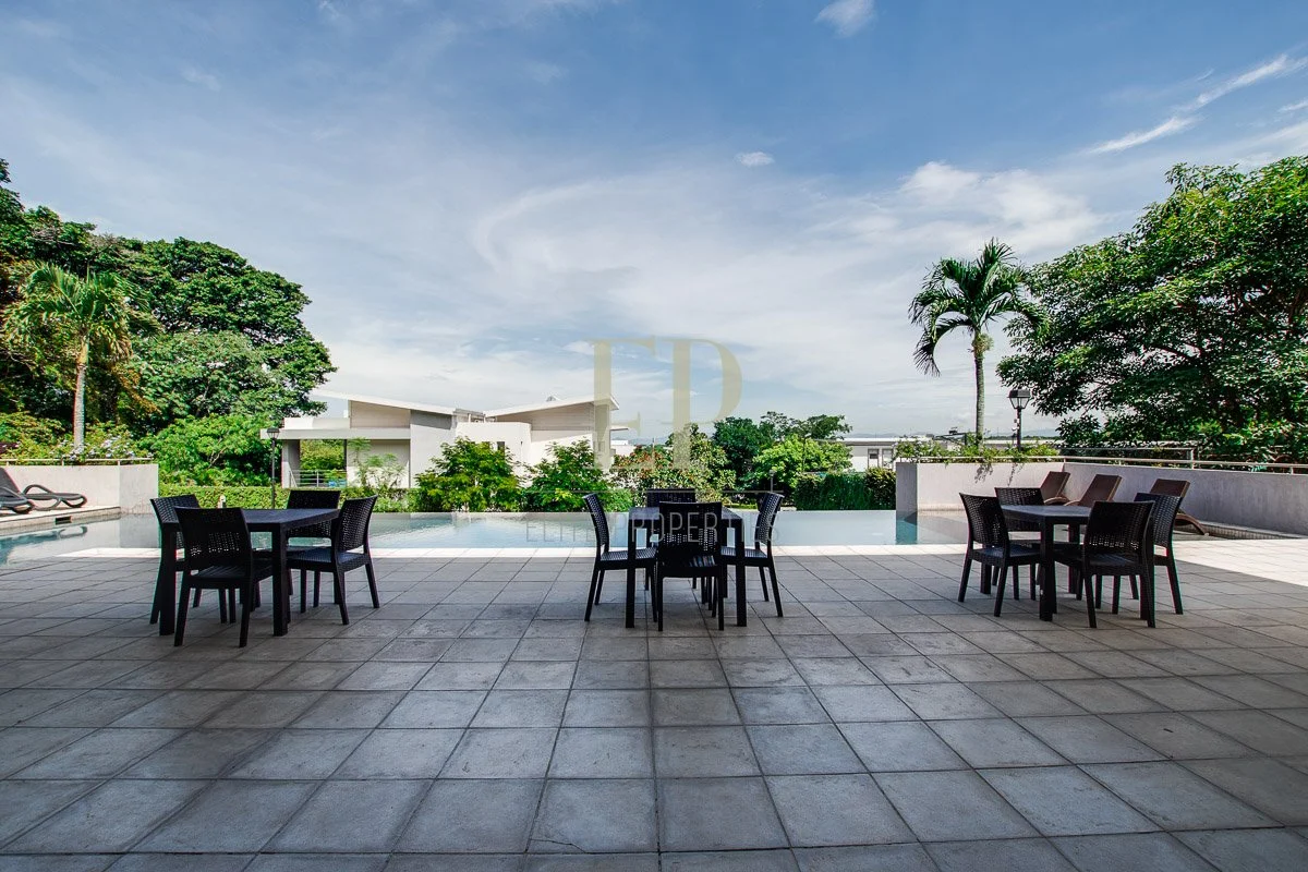 Outdoor patio with black tables and chairs, pool, lounge chairs, and lush greenery under a blue sky.