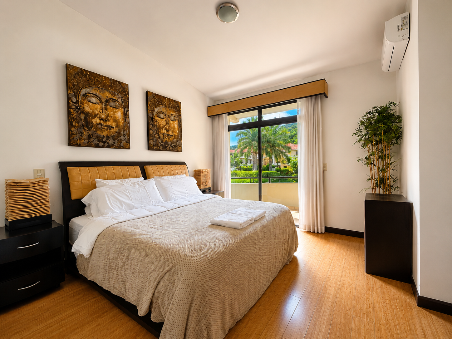 A bedroom with a large bed, white bedding, and a tan headboard. Two nightstands with black drawers and wicker lamps. Two framed art pieces of Buddha faces above the bed. A sliding glass door with white curtains leads to a balcony with tropical trees 