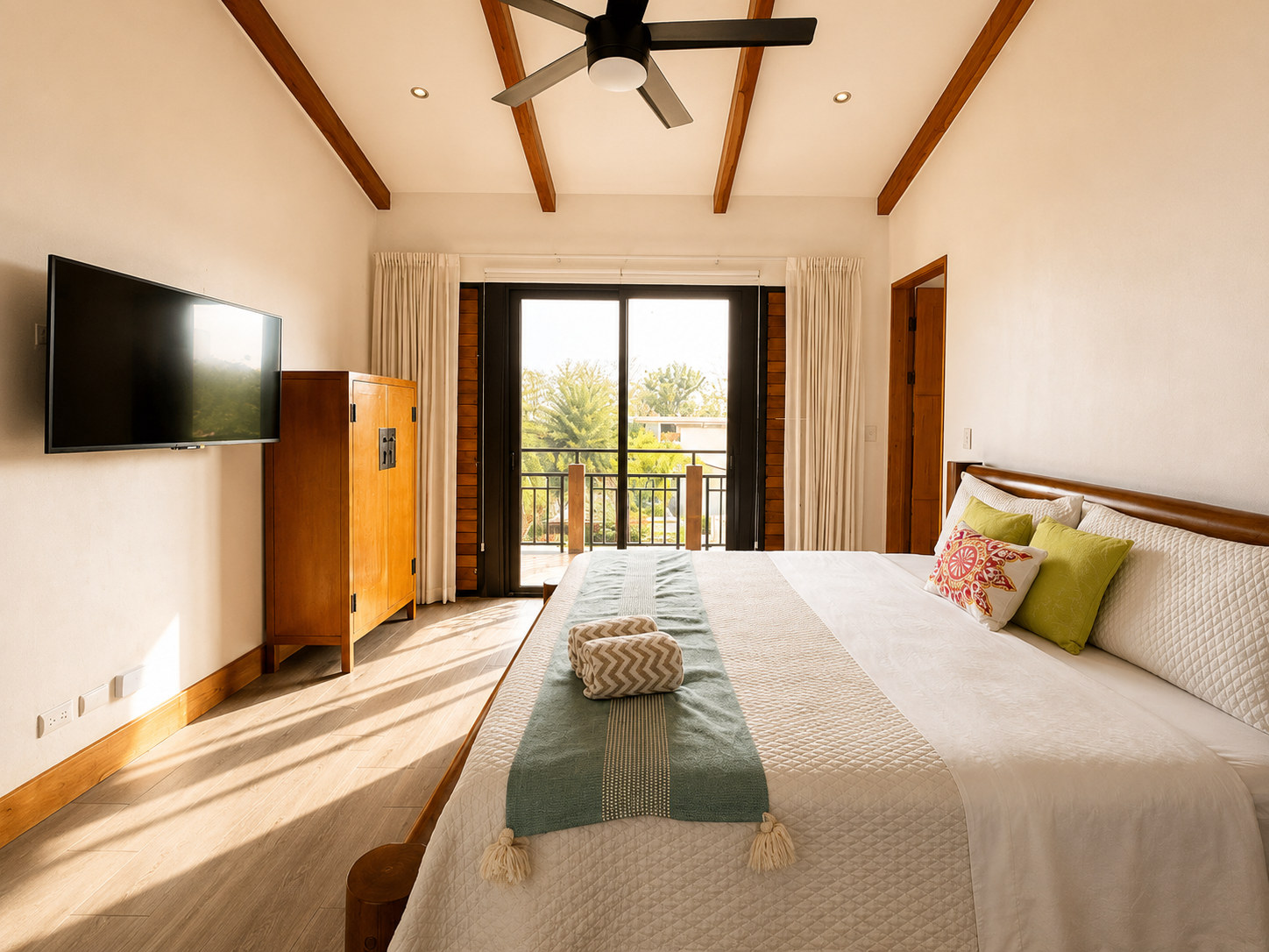 Hotel room with a large bed, colorful pillows, and a balcony with a view of trees and neighborhood.