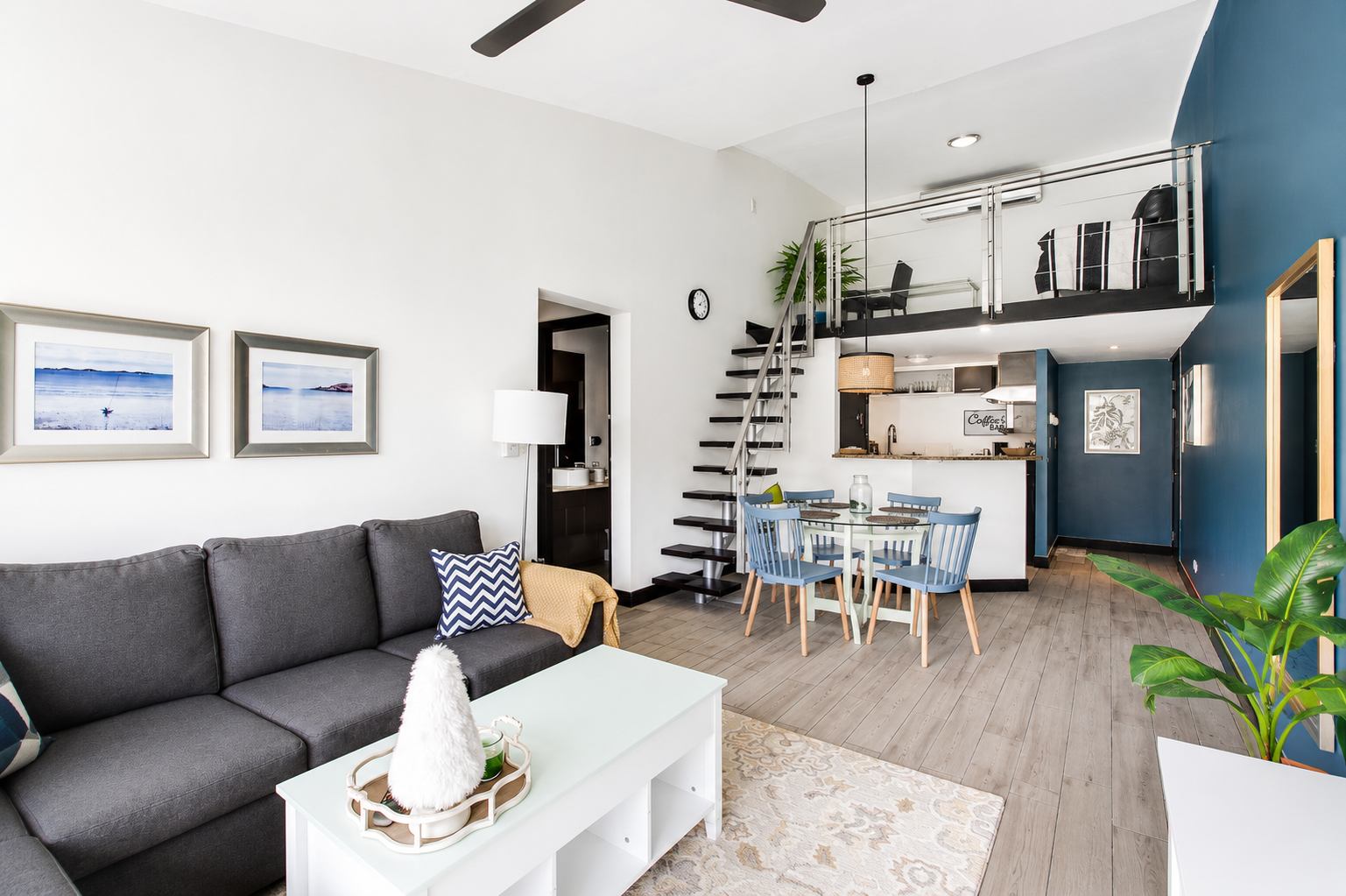 An open-concept living room and dining area with modern furniture, including a gray sofa with patterned pillows, a white coffee table with a decorative tray, a light wood dining table with blue chairs, and a staircase leading to a loft with an additi