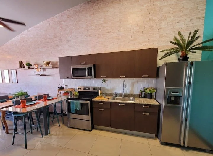 Open-concept kitchen with dark brown cabinets, stainless steel appliances including a refrigerator, stove, and microwave. There's a beige tiled backsplash, a sink beneath wall cabinets, and a countertop with plants and bottles. Adjacent to the kitche